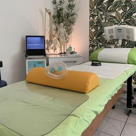 Behandlungsliege mit grüner Auflage, Beatmungsmaske, Gerät über dem Kopfbereich, Laptop und Anatomiemodellen im Hintergrund.