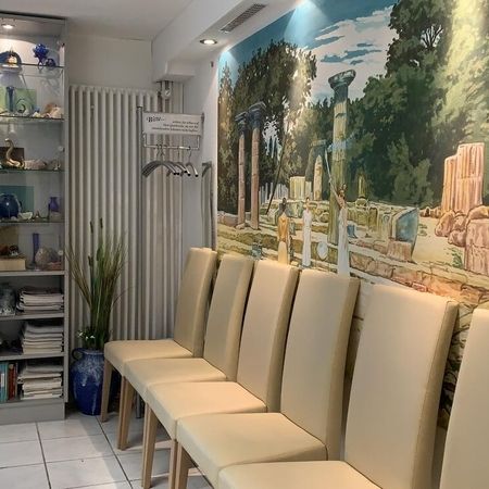 Wartezimmer mit beigen Stühlen, Glasvitrine und Wandbild mit antikem Motiv.