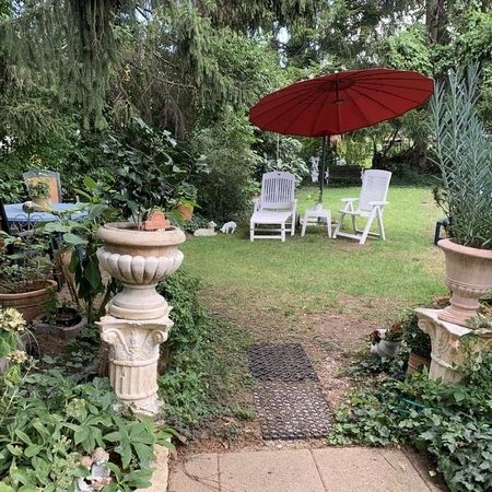 Garten mit weißen Gartenstühlen, rotem Sonnenschirm und vielen Pflanzen.