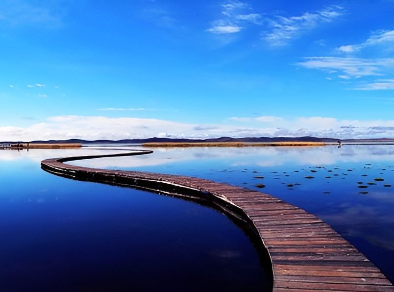 Geschwungener Holzsteg auf ruhigem See mit Spiegelung des Himmels - beeindruckende Landschaft mit weitem Horizont und sanften Hügeln im Hintergrund
