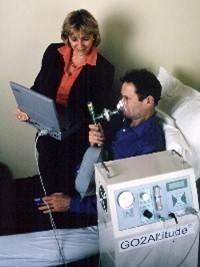 Ein Mann liegt im Bett und atmet durch eine Maske, daneben eine Frau mit Laptop, vor einem Gerät mit der Aufschrift GO2Altitude